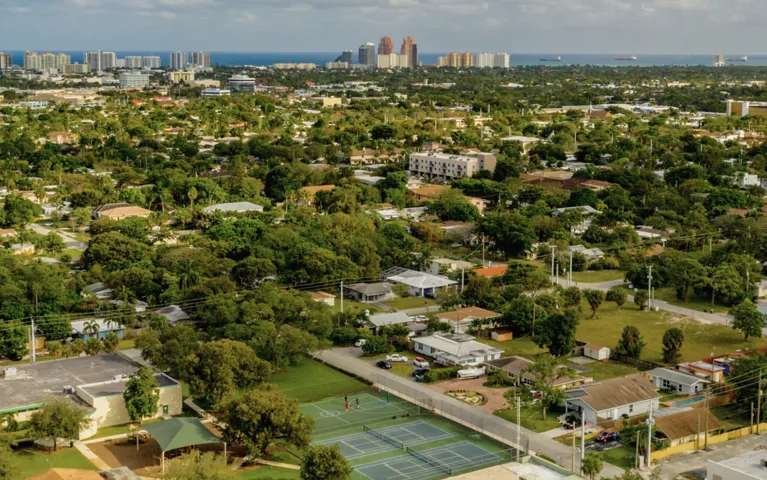 Aerial view of a suburban neighborhood with tennis courts, dense greenery, residential houses, and distant high-rise buildings near the coastline.
