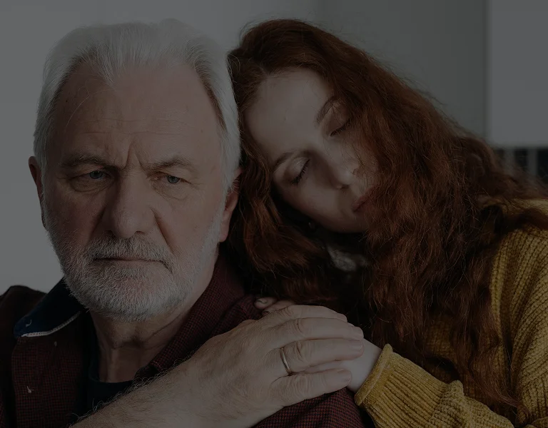Older man with white hair and beard being comforted by younger woman with long red hair and yellow sweater.
