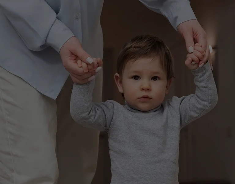 Toddler in grey shirt holding hands with adult wearing light blue shirt and white pants.