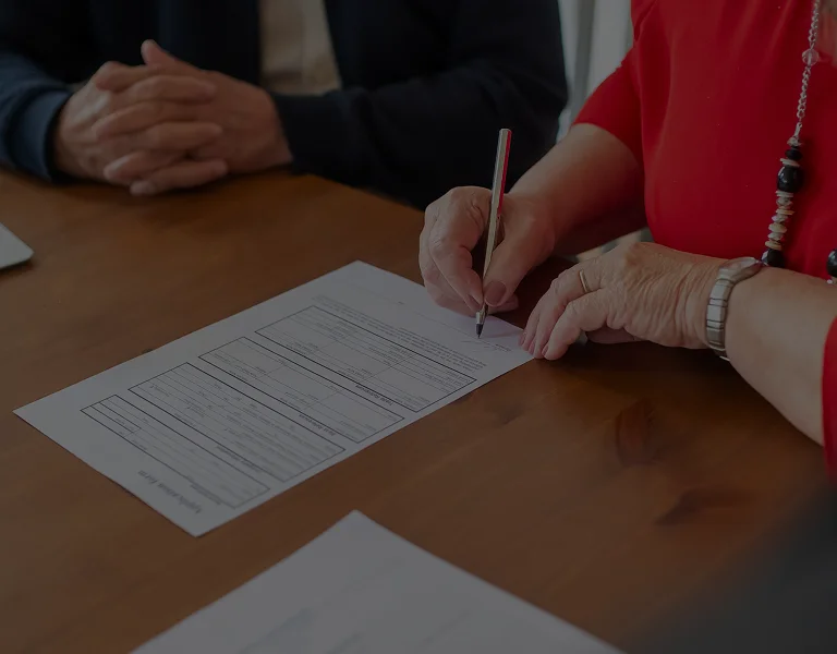 Person in red attire signing a document on a wooden table, another person with folded hands nearby.