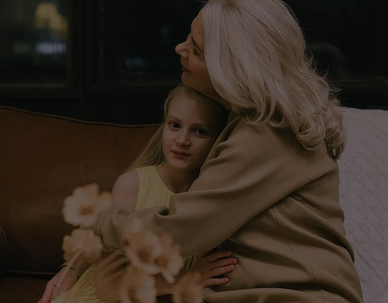 Elderly woman with gray hair hugging young girl in yellow dress on brown sofa.