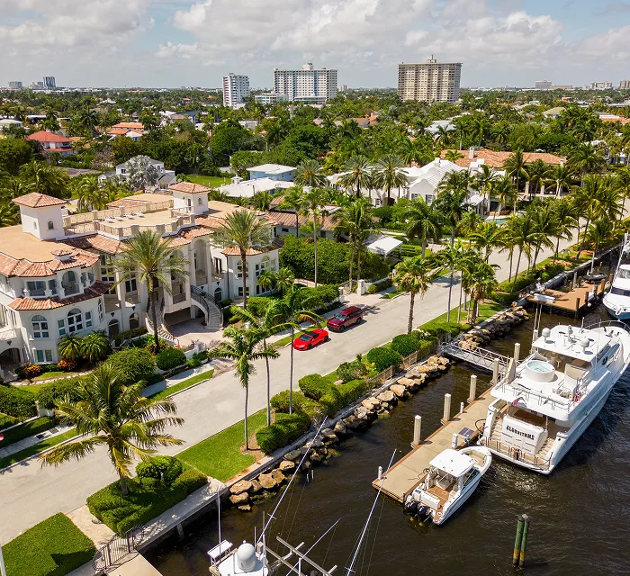 Aerial view of waterfront luxury homes, palm trees, parked cars, and yachts docked along a marina.