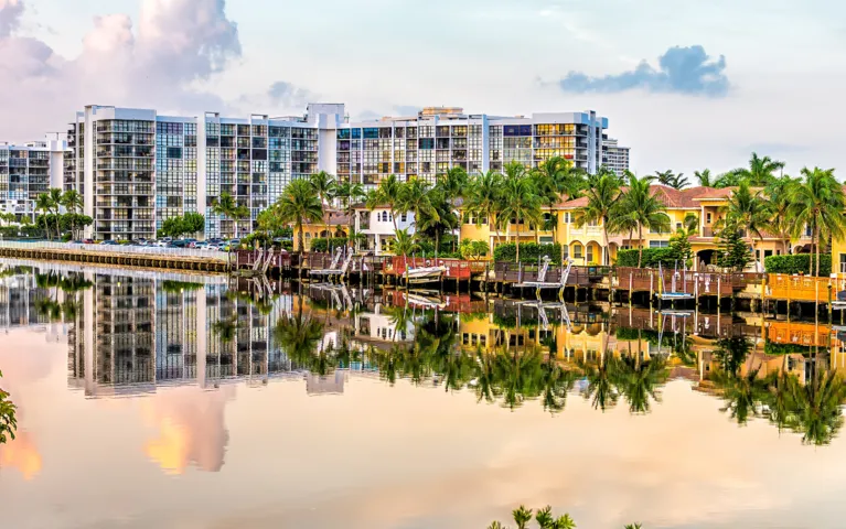 Waterfront residential buildings and palm trees reflected in calm canal water under a pastel sky.