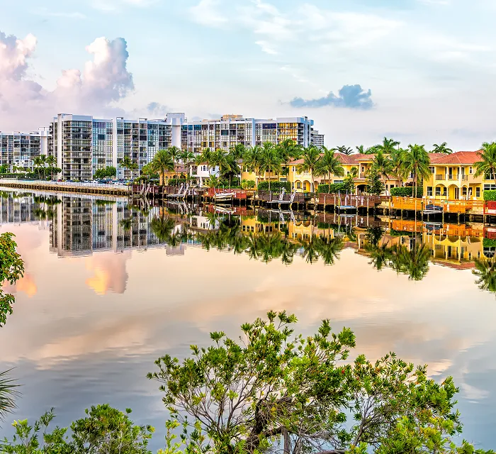 Waterfront residential area with modern buildings, palm trees, and calm reflective water.