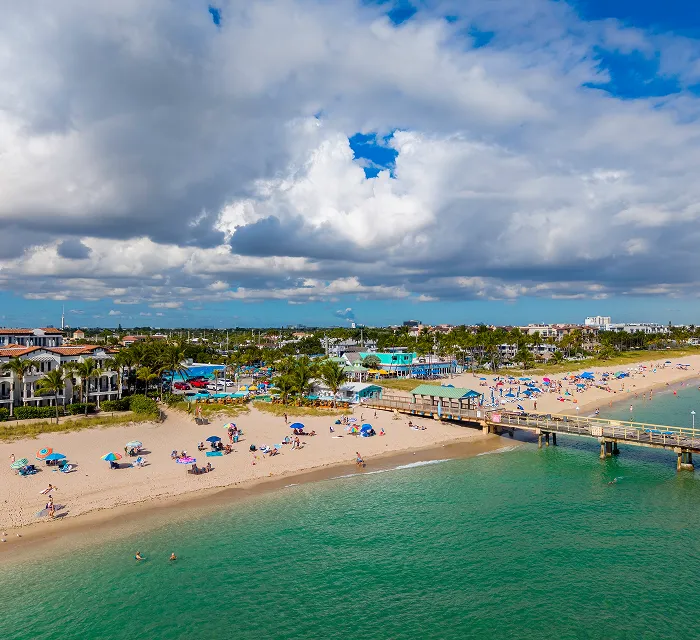 Aerial view of a sandy beach with colorful umbrellas, a wooden pier, turquoise water, and partly cloudy sky.