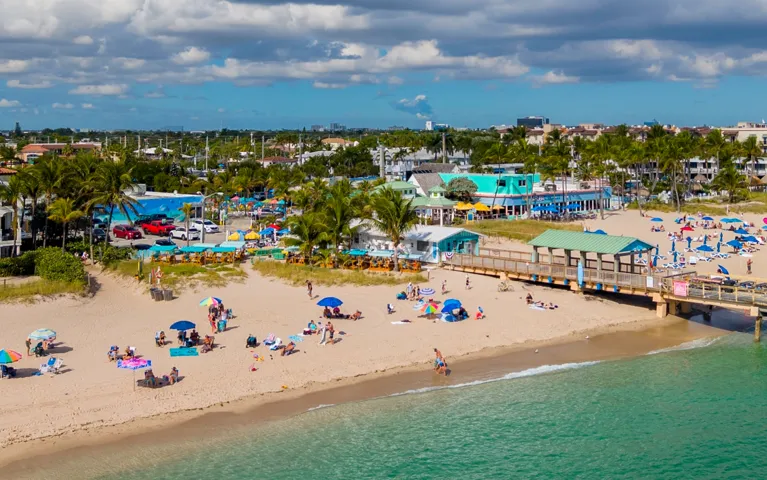 A sunny beach scene with people under colorful umbrellas, a wooden pier, turquoise water, and palm trees.