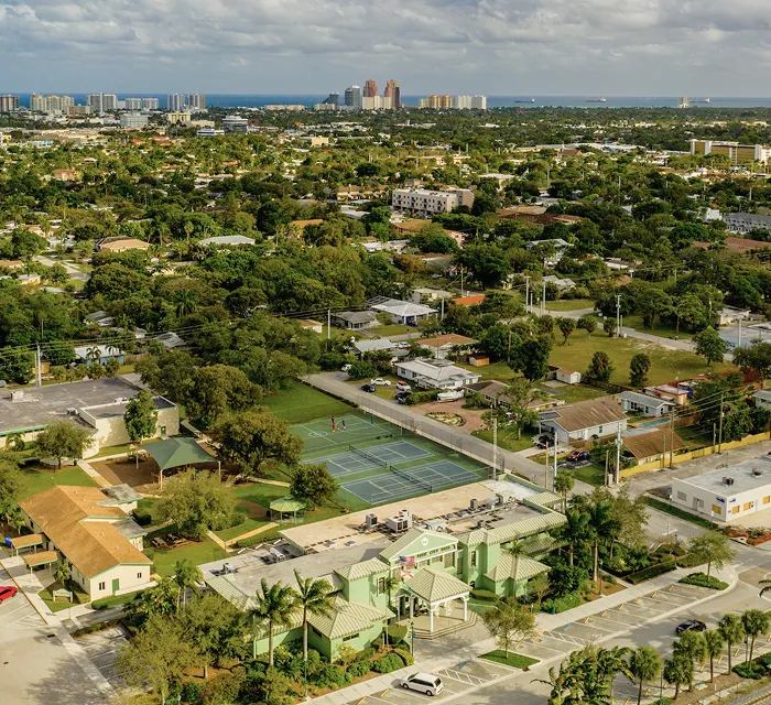 Aerial view of a suburban neighborhood with tennis courts, greenery, and distant high-rise buildings.