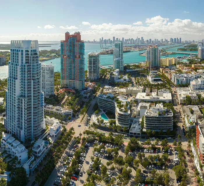Aerial view of a coastal city with high-rise buildings, waterways, and parking lots under a partly cloudy sky.