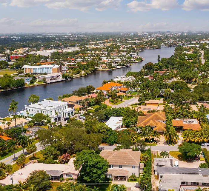 Aerial view of a residential neighborhood with a river running through, featuring houses, greenery, and partly cloudy sky.