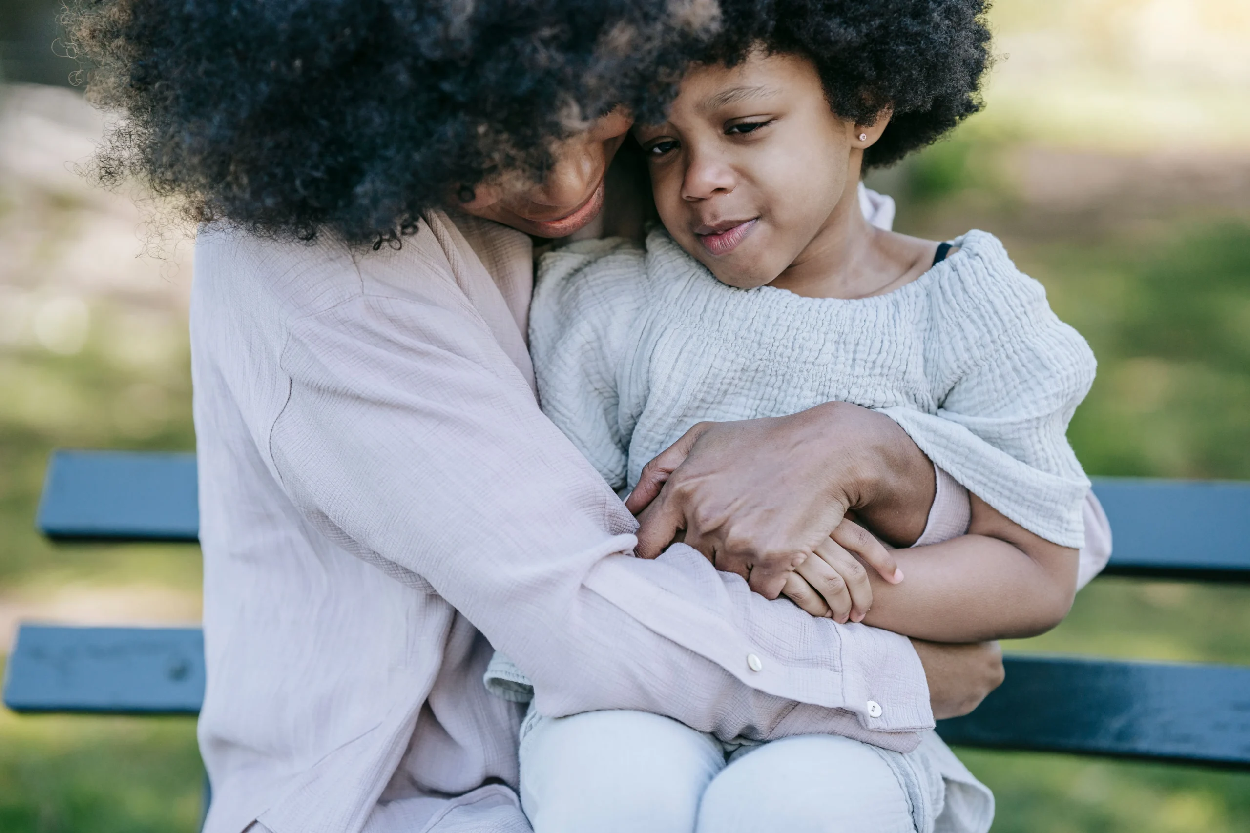 African American woman comforting a distressed young girl on a park bench outdoors.