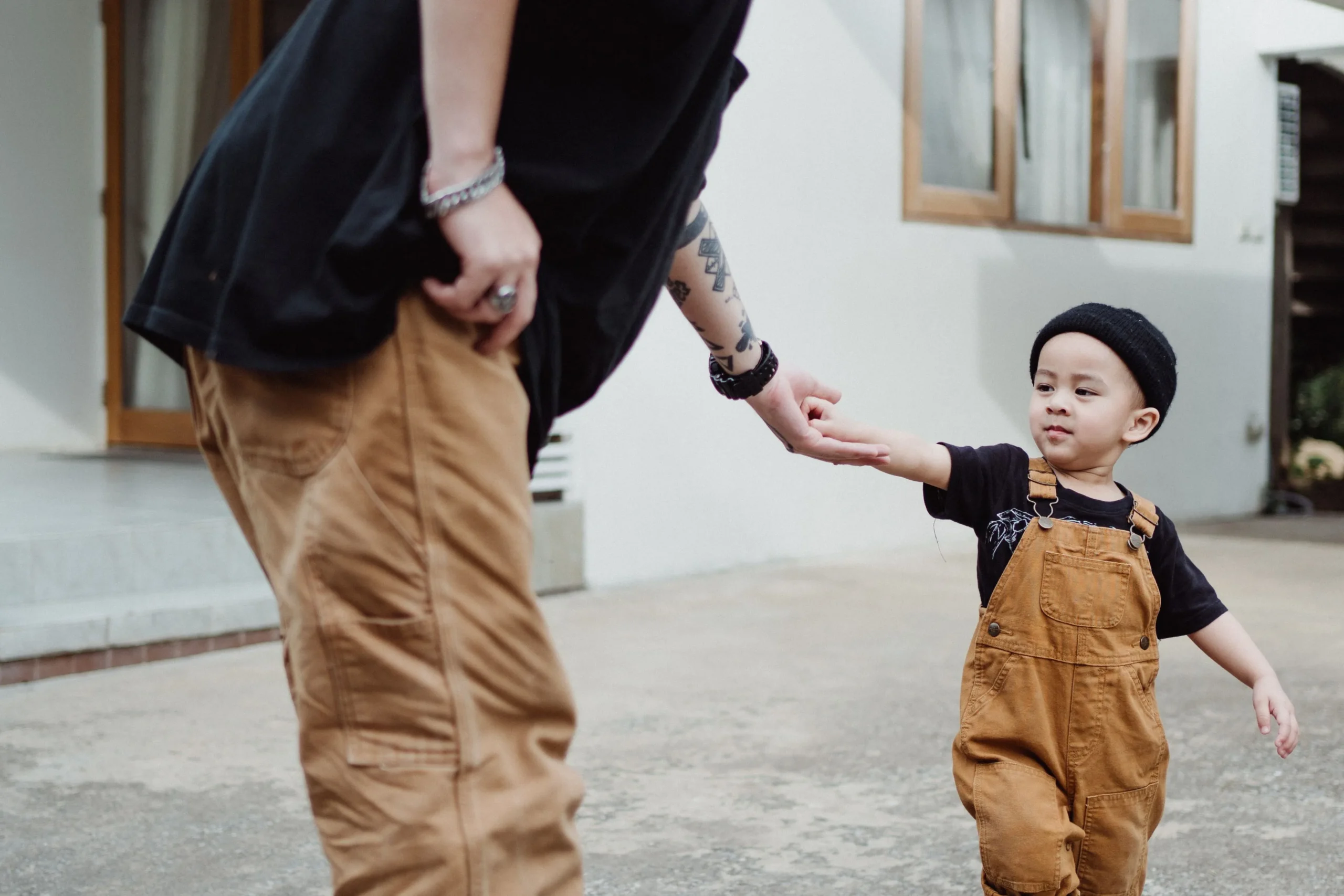 Young child in brown overalls and black beanie holding hands with tattooed adult in tan pants outdoors.