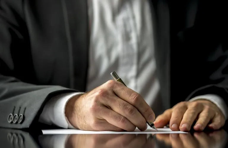 Close-up of a person in a dark suit writing on white paper with a pen.